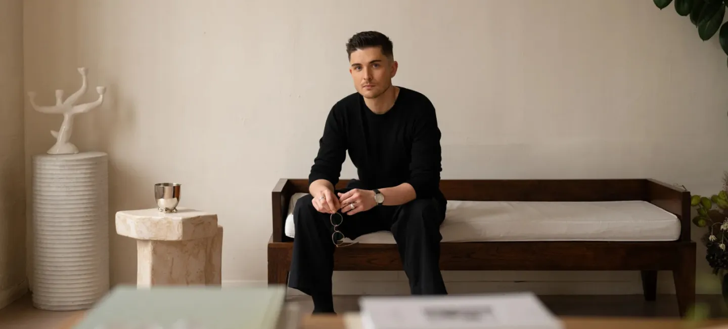 Mężczyzna w czarnym stroju siedzący na drewnianej sofie w jasnym, minimalistycznym wnętrzu