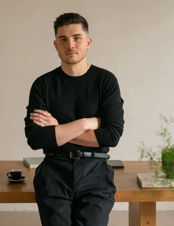 Mężczyzna w czarnym swetrze ze skrzyżowanymi rękami, oparty o drewniany stół z filiżanką espresso, książkami i laptopem